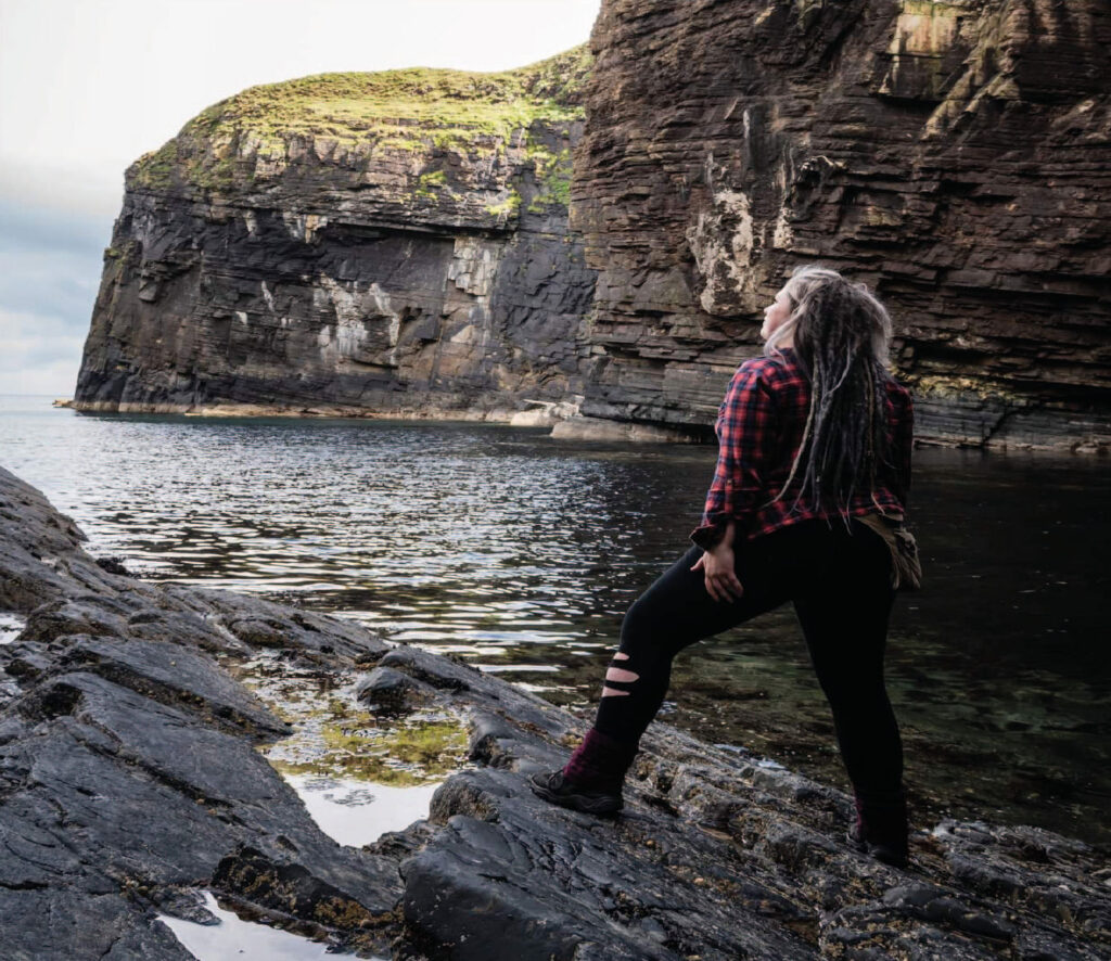 An image showing a women standing on stone by the water looking into the distance