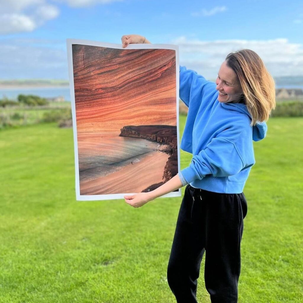 A image showing Lisa Poulsen holding a paper painting standing on a green grassy field