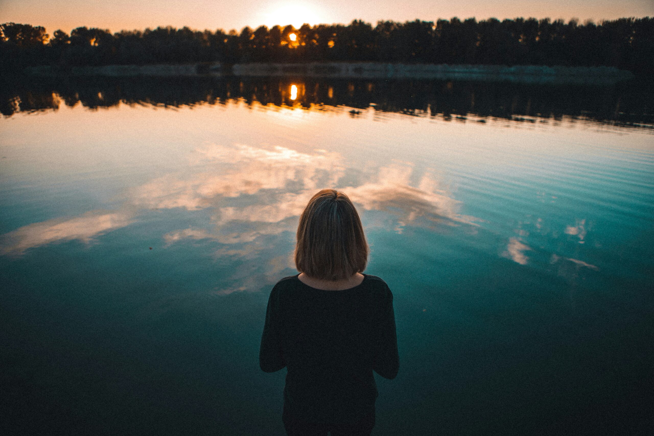 a women staring away from the camera looking at water with a treeline and sunset in the backdrop.